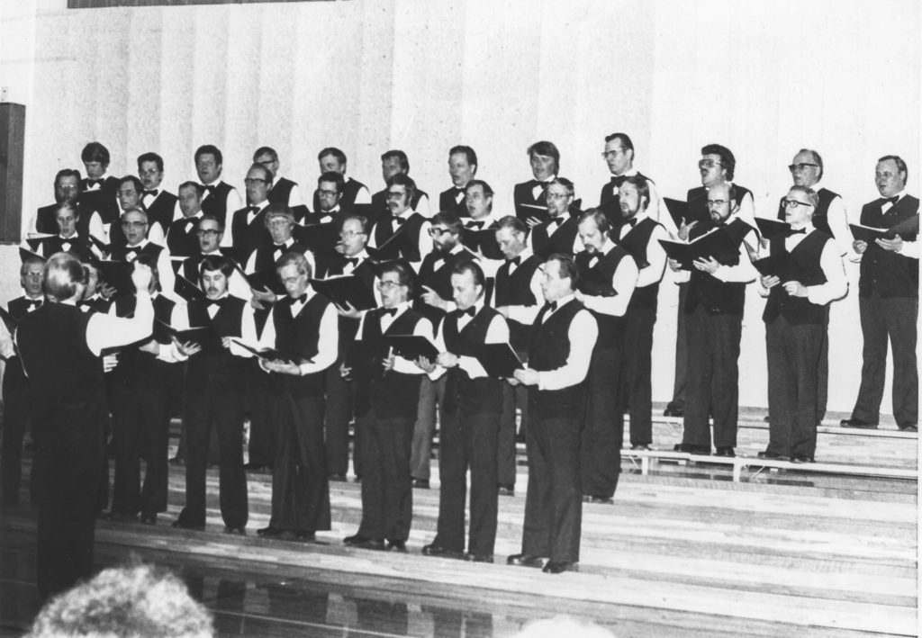 Mustavalkoinen kuva mieskuorosta laulamassa liivipuvuissaan.
