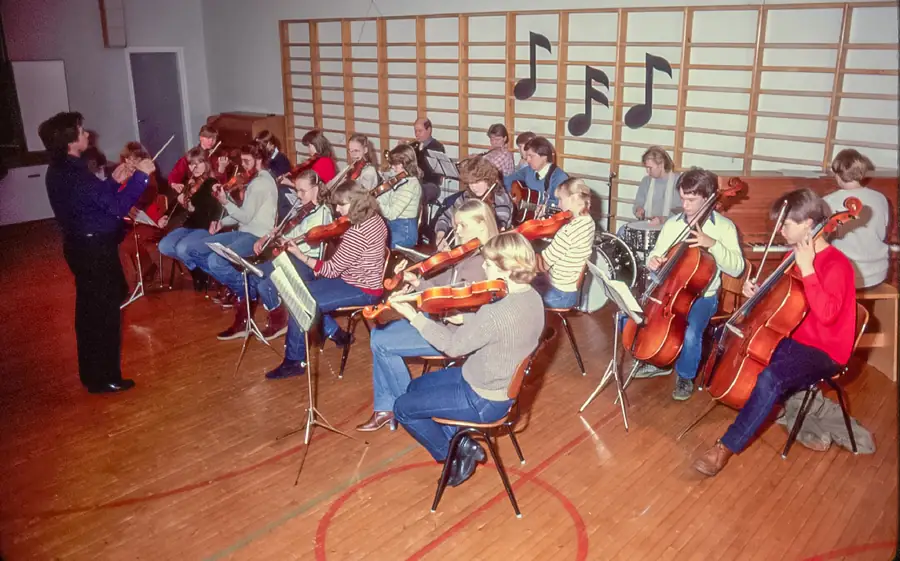 Eri instrumenttejä soittava orkesteri esiintyy jumppasalissa opettajan johdolla. Takana oleviin puolapuihin on kiinnitetty kolme isoa pahvista nuottia.