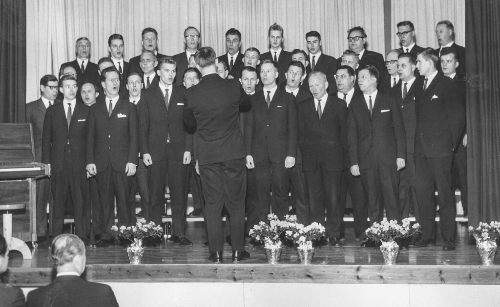 Kuvassa noin 30-jäseninen mieskuoro laulamassa. Kaikilla tumma puku päällä.