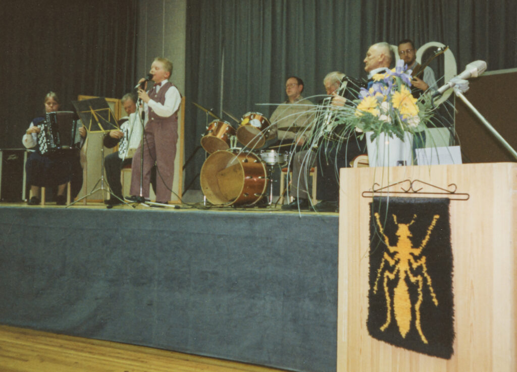 Lavalla seisoo nuori poika laulusolistina, säestämässä yhtye, jossa on harmonikkoja, kitara ja rummut. Etualalla lavan sivustalla on pieni ryijy, jossa on Multian vaakuna, eli keltainen muurahainen mustalla pohjalla.