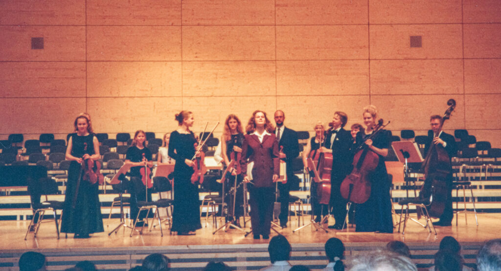 Esiintymislavalla seisoo nuoria viulun, sellon ja komtrabasson soittajia. Orkesterin johtaja on kääntynyt yleisöön päin vastaanottamaan aplodeja.