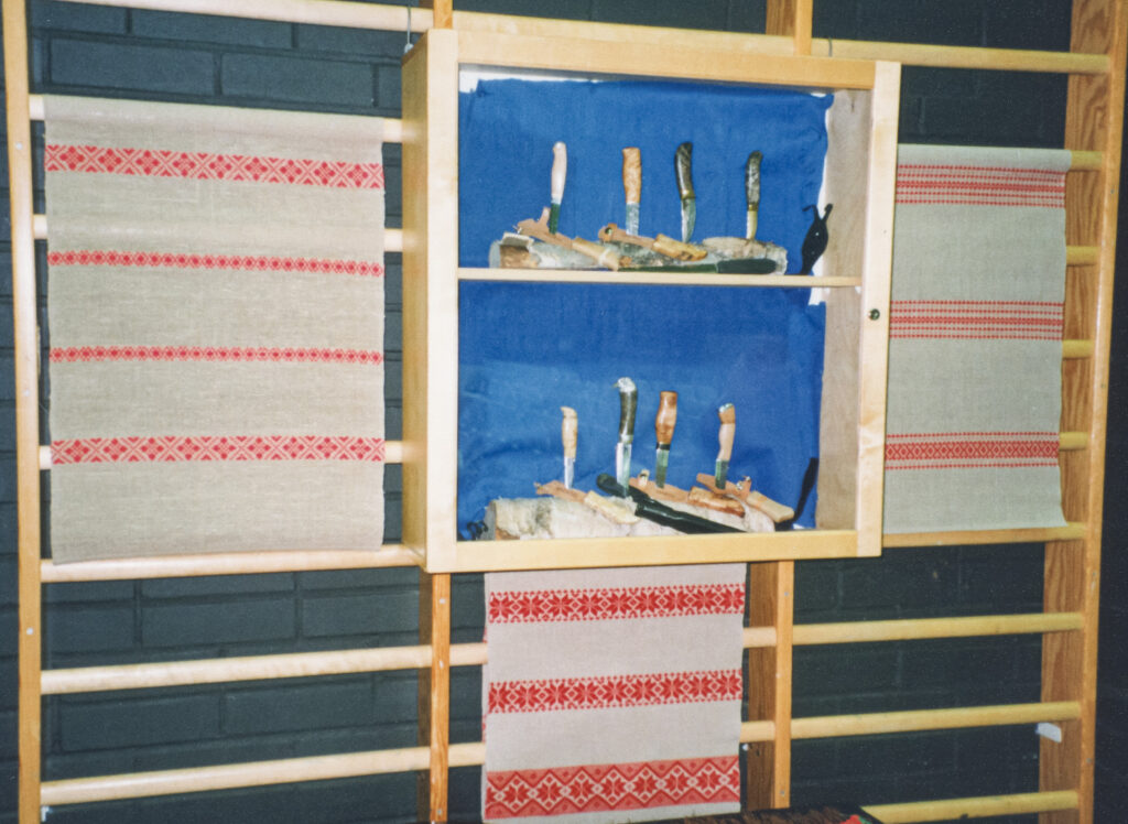 Puolapuille on ripustettu kangaspuilla kudottuja liinoja. Keskellä on hylly, jossa on taontakurssilla valmistettuja puukkoja.
