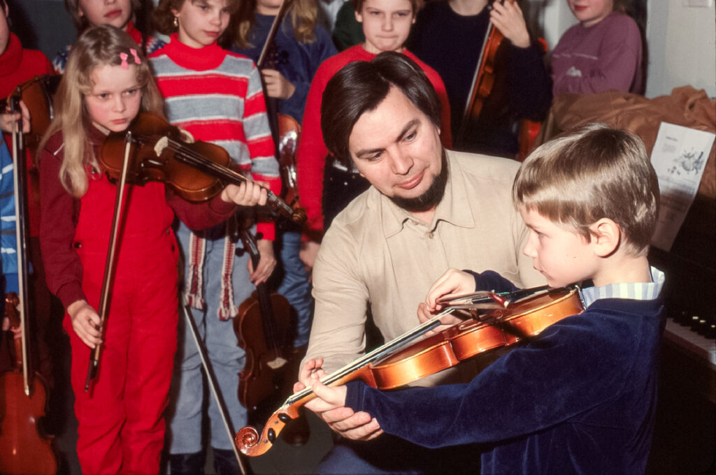 Etualalla ystävällisesti hymyilevä mies ohjaa viulua soittavan pikkupojan käsien asentoa. Taustalla odottaa vuoroaan kahdeksan lasta viulut käsissään.