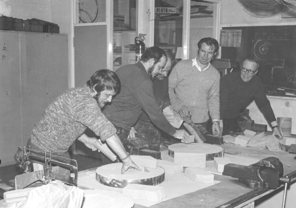 Mustavalkoisessa kuvassa on viisi miestä työpöydän ääressä rakentamassa uunin osia metallityökerhossa.
