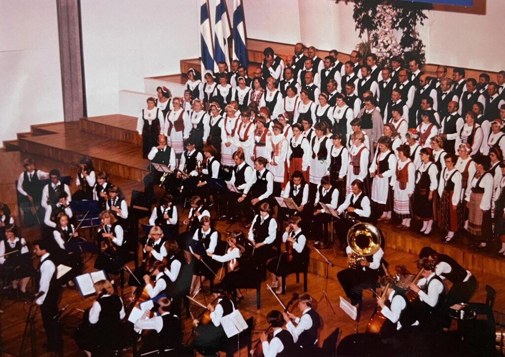 Esiintymässä on kymmeniä kuorolaisia kansallispuvuissa. Kuoron edessä istuu orkesteri, jossa on nuoria viulun, sellon ja puhaltimien soittajia. Taustalla on kolme Suomen lippua.