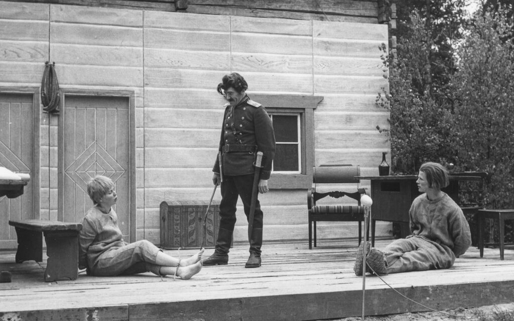 Yksi näyttelijä seisoo sotilaspuvussa, vieressä istuu maassa kaksi näyttelijää sidottuina vangeiksi.