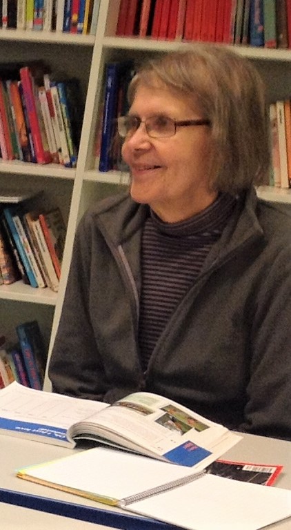 Silmälasipäinen nainen hymyilee istuessaan kirjan ääressä.