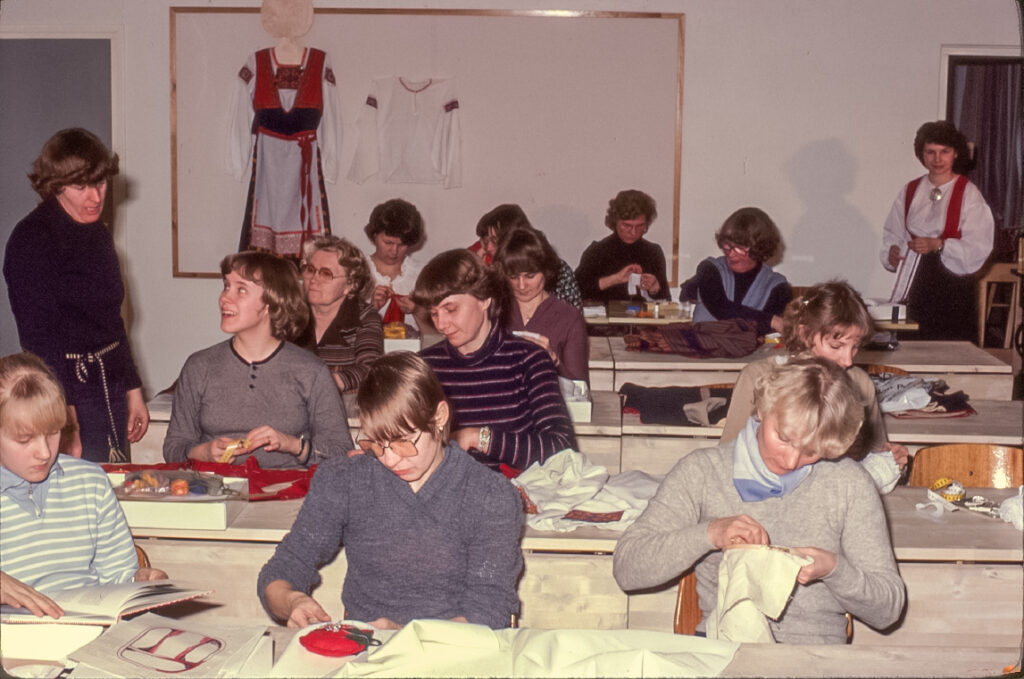 12 naista istuu pöytien ääressä ompelemassa käsin. Opettaja seisoo pöydän vieressä keskustelemassa opiskelijan kanssa. Taustalla näkyy seinälle ripustettu kansallispuku ja yksi opiskelija pukeutuneena kansallispukuun.