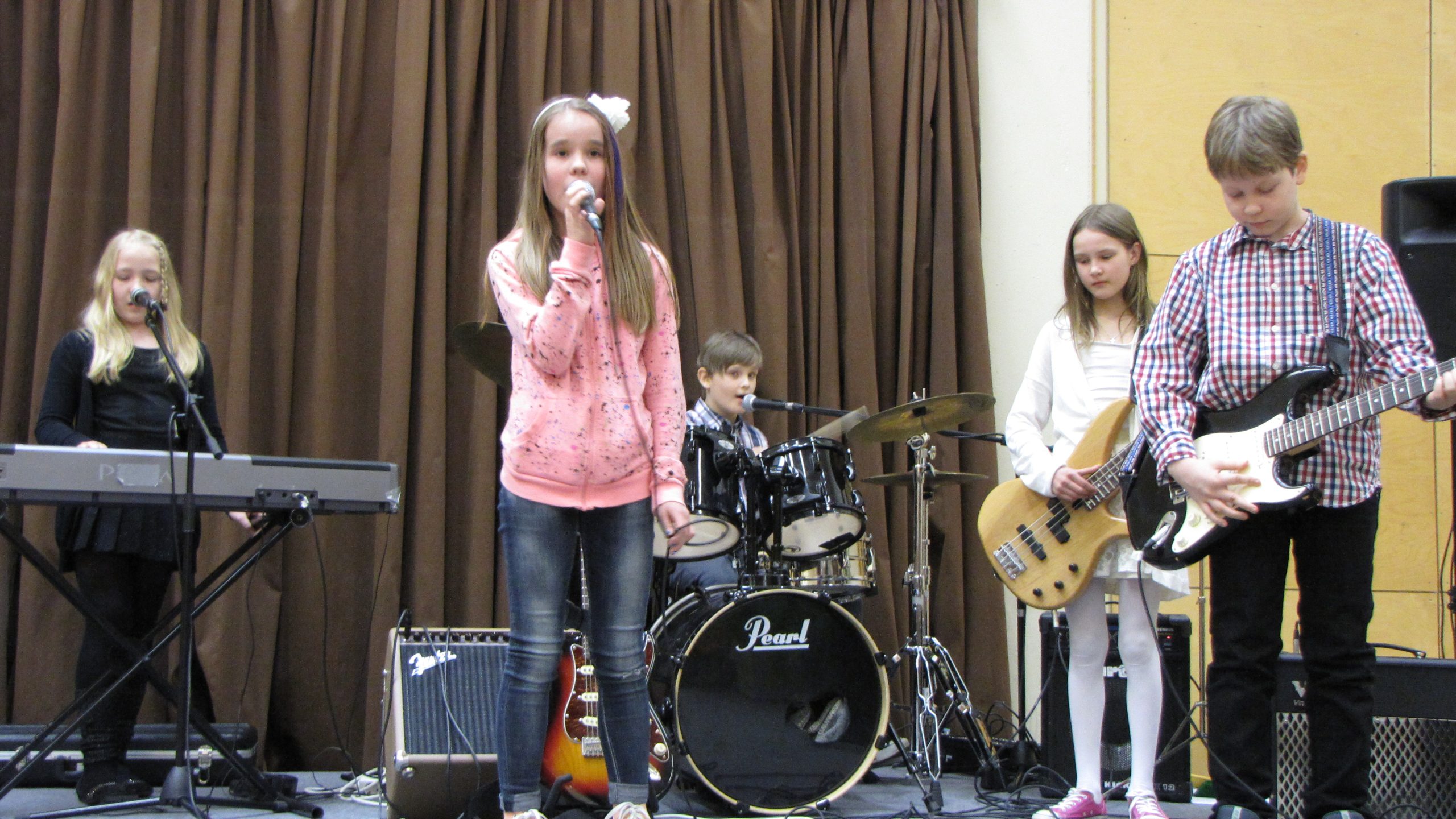 Alakouluikäiset kaksi poikaa ja kolme tyttöä esittävät bändinä kappaletta, yksi tytöistä laulaa.