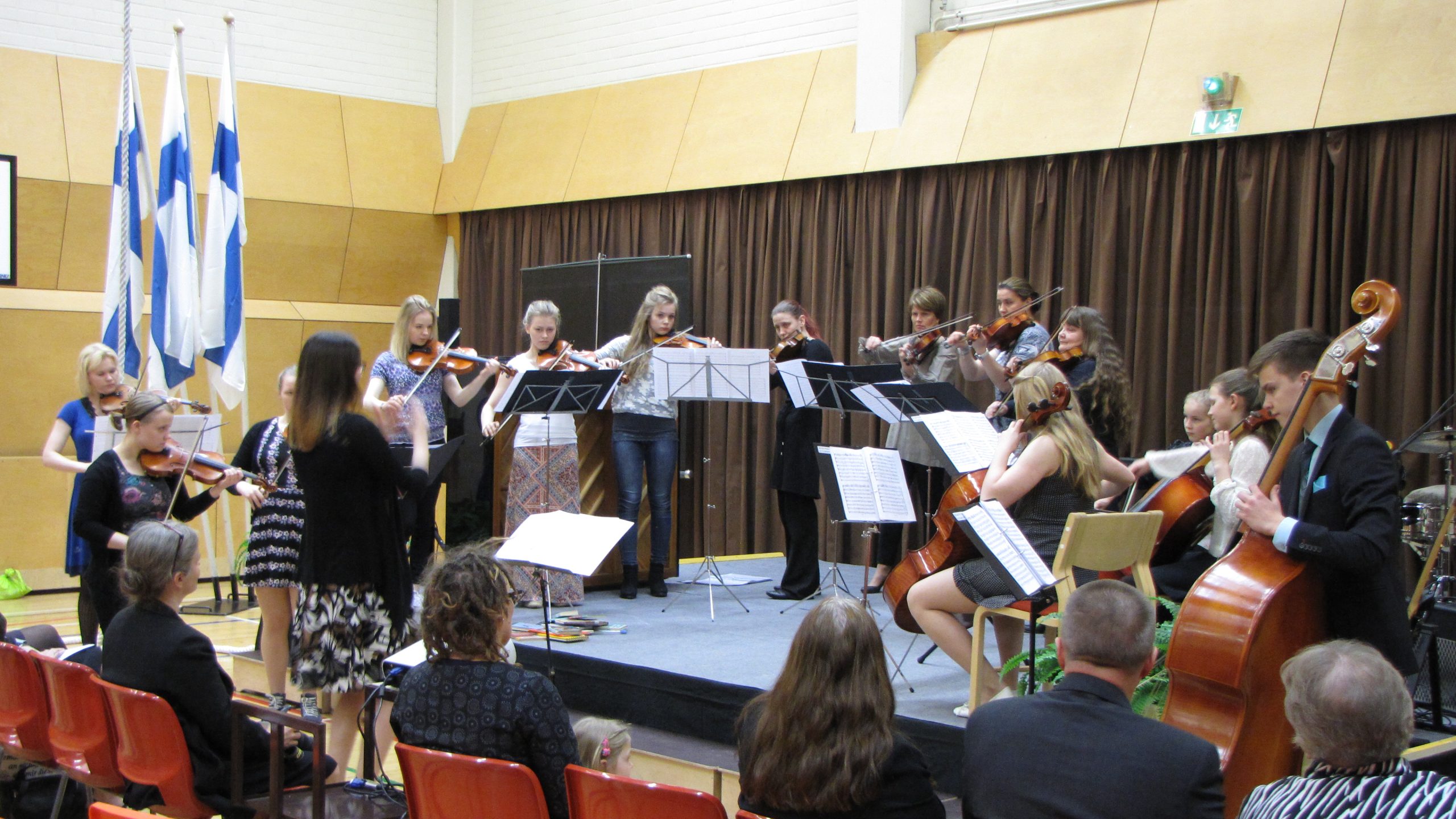 Nuorista koostuva viulu- ja sello-orkesteri esiintyy, nainen johtaa soittoa.