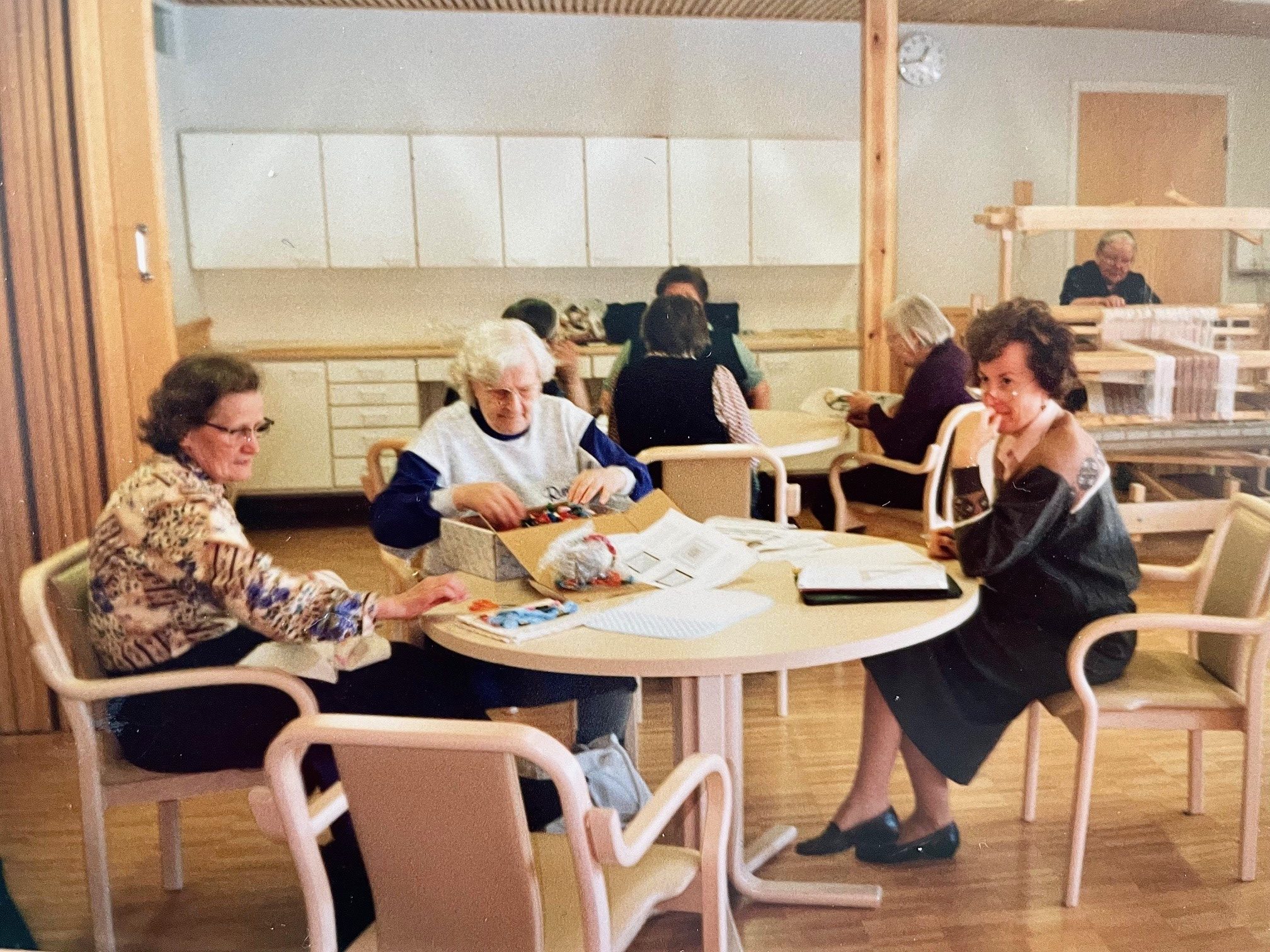 Ryhmä naisia istuu kahden pyöreän pöydän ympärillä tekemässä käsitöitä.