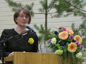 Kuvassa nainen puhujapöntössä värikkään kukka-asetelman vieressä.