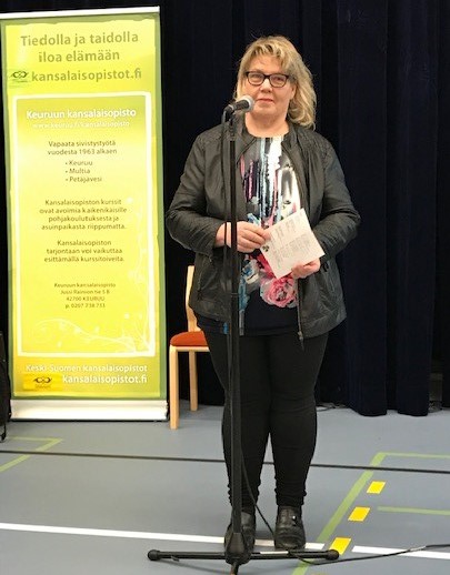 Nainen seisoo mikrofonin edessä paperilappu kädessään. Takana keltainen banneri, jossa lukee Tiedolla ja taidolla iloa elämään, kansalaisopistot.fi.