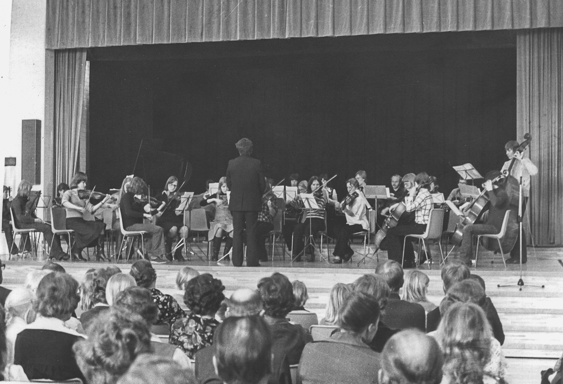 Vanhassa kuvassa iso orkesteri esiintyy juhlasalin lavalla., etualalla näkyy yleisöä.