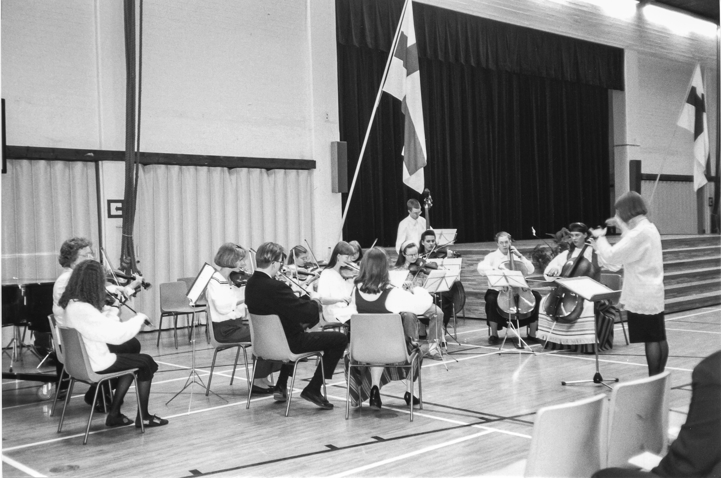 Orkesteri esiintyy juhlasalissa lavan vieressä permannolla opettajansa johdolla. Taustalla näkyy kaksi Suomen lippua.