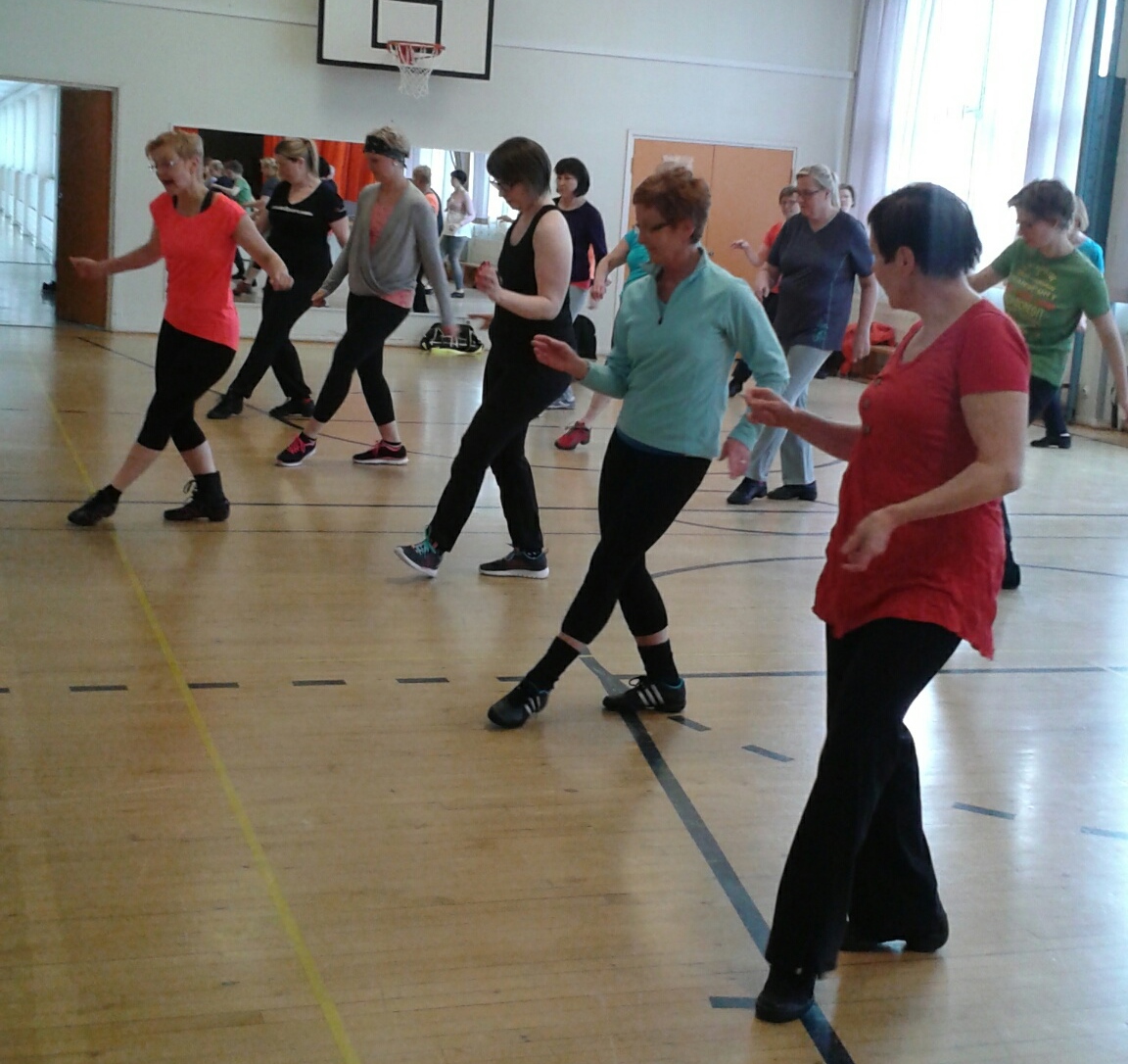 Ryhmä naisia koulun jumppasalissa opettelee tanssia opettajan johdolla. Jokaisella on toinen jalka työnnettynä eteen.