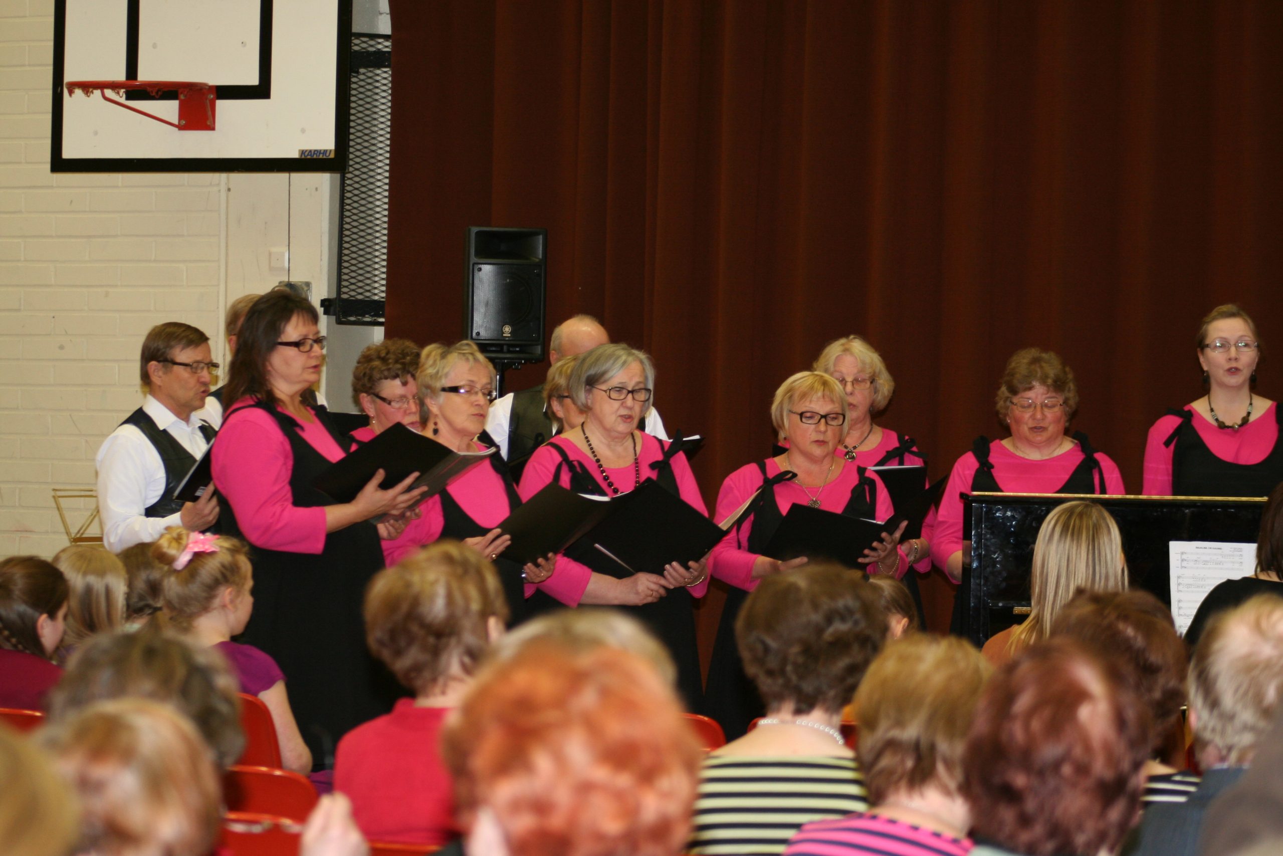 Yhdeksän naista pinkeissä paidoissa ja kolme miestä valkoisissa paidoissa esittää kuorolaulua yleisölle.