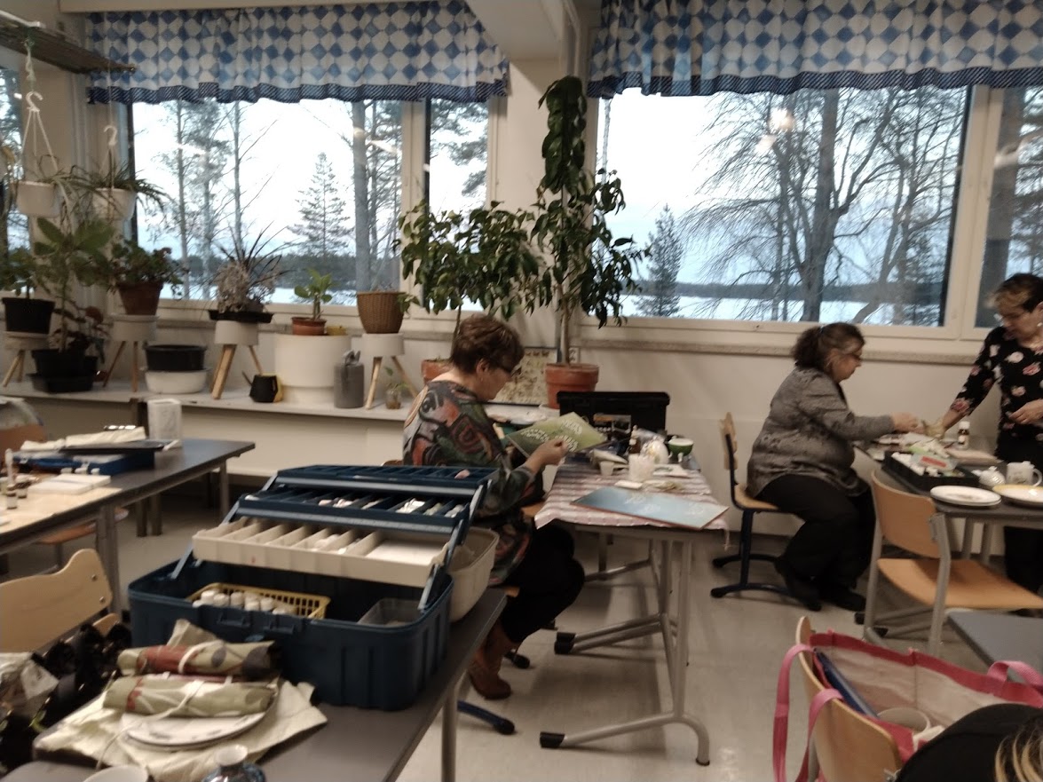 Kolme naista tekee pöytien ääressä posliinitöitä, ympärillä on paljon tavaraa. Ikkunoista näkyy talviselle järvelle ja ikkunalaudalla on monta kasvia.