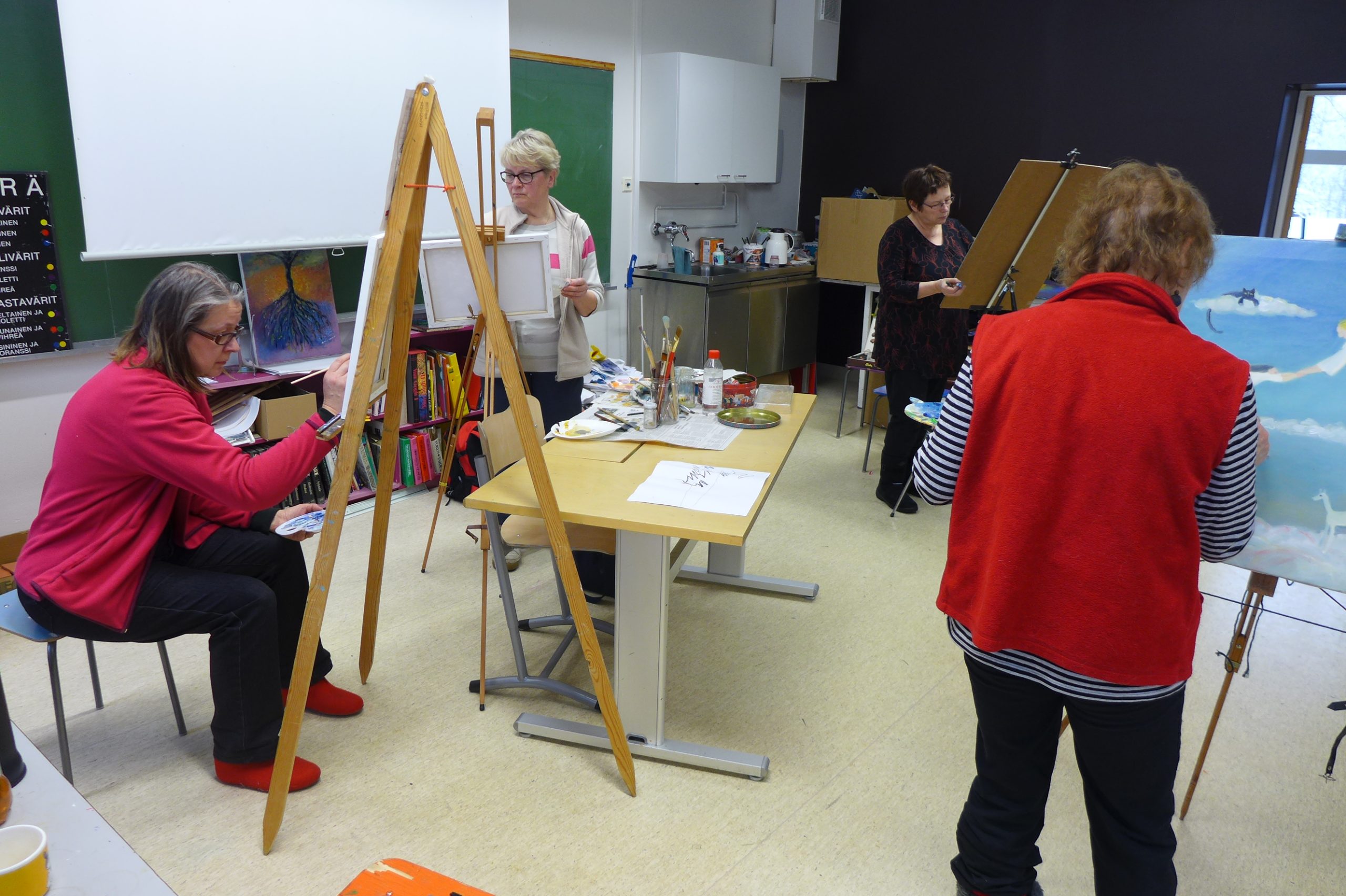 Luokkahuoneessa kolme naista seisoo ja yksi istuu telineidensä ääressä maalaamassa taulua.