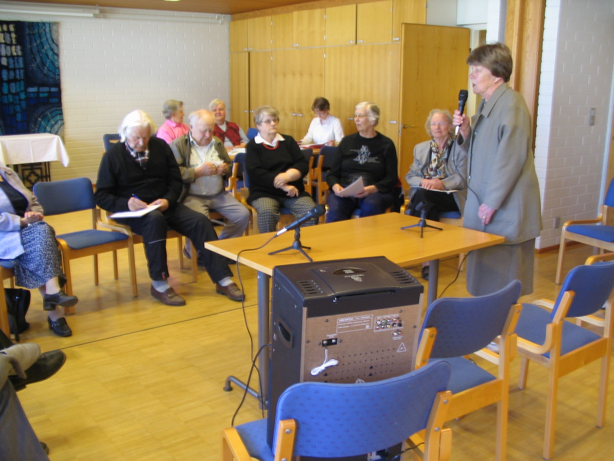 Ryhmä senioreita istuu tuoleilla puoliympyrässä, etualalla nainen puhuu mikrofoniin. Taustalla kolme ihmistä pöydän ääressä.
