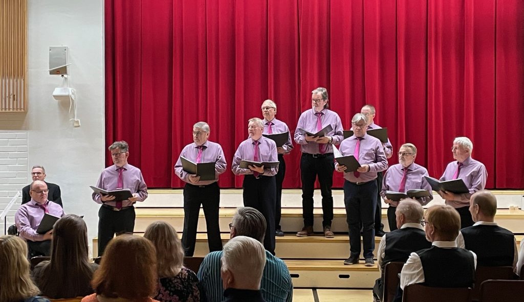 Koulun juhlasalissa kymmenen miestä esittää kuorolaulua yleisölle.