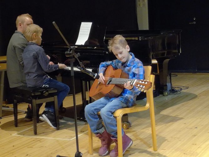 Poika istuu tuolilla soittamassa akustista kitaraa, taustalla toinen poika opettajansa kanssa soittaa pianoa.