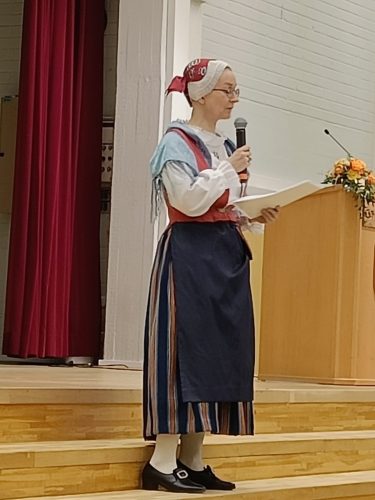 Kansallispukuinen nainen puhuu mikrofoniin yleisölle.