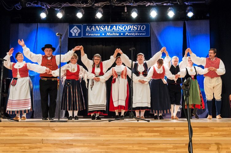 Kansallispukuihin pukeutuneet tanhupiiriläiset seisovat lavalla käsi kädessä kiittämässä yleisöä.