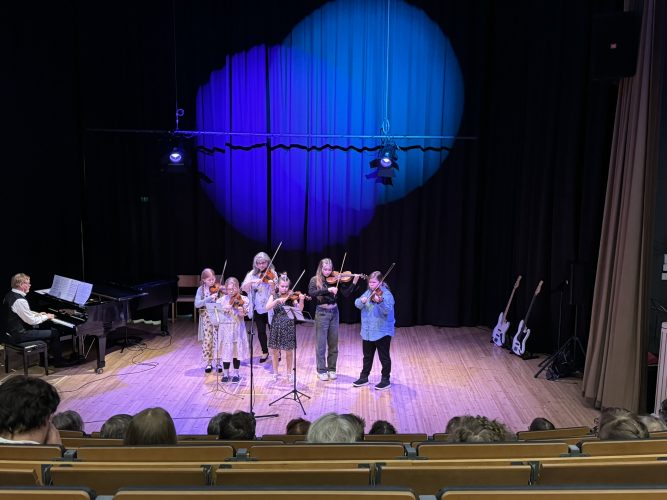 Viisi koulutyttöä opettajansa kanssa esittävät viulunsoittoa. Mies soittaa taustalla pianoa.