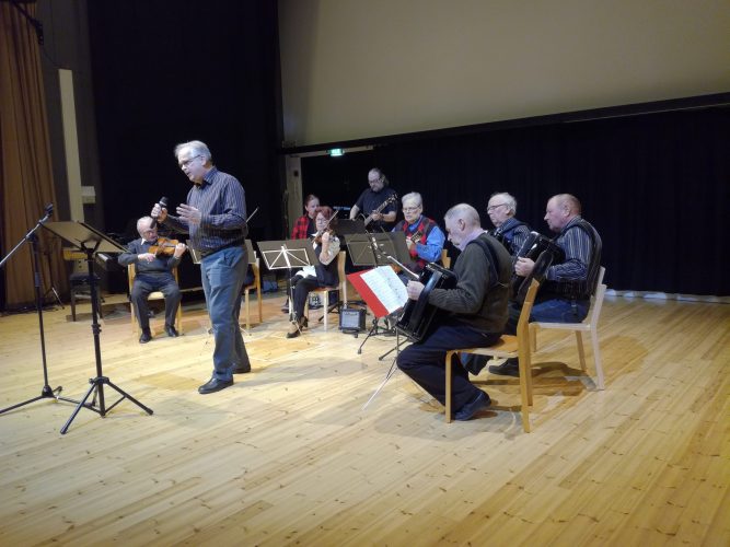 Mies seisoo ja laulaa mikrofoniin ja taustalla pelimannit istuvat ja soittavat eri instrumentteja.