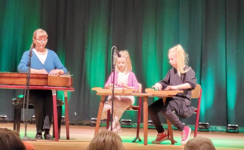 Kaksi lasta ja yksi aikuinen soittaa kanteletta esiintymislavalla.