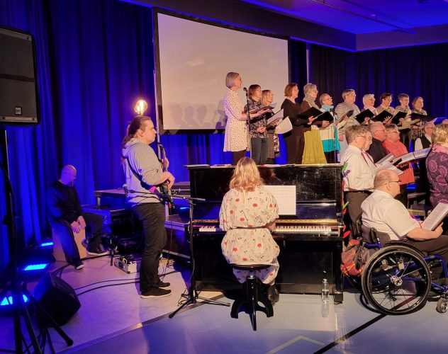 Etualalla mies soittaa bassoa ja nainen pianoa, taustalla kuoro laulamassa.