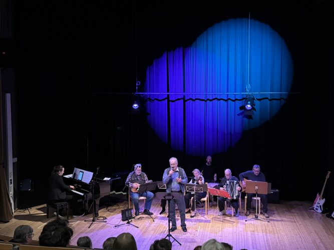 Kuusi pelimannisoittajaa istuu soittamassa, edessä mies laulaa mikrofoniin ja taustalla toinen mies soittaa pianoa.