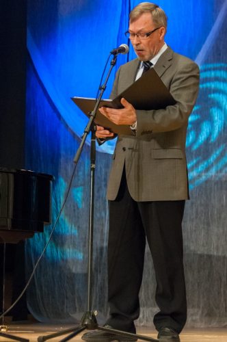 Mies harmaa puvuntakki päällään ja pitäen nuottikansiota käsissään laulaa mikrofoniin.