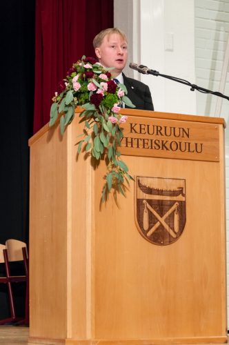 Mies pitää puhetta. Puhujapöntössä lukee Keuruun yhteiskoulu ja siinä on kukka-asetelma.