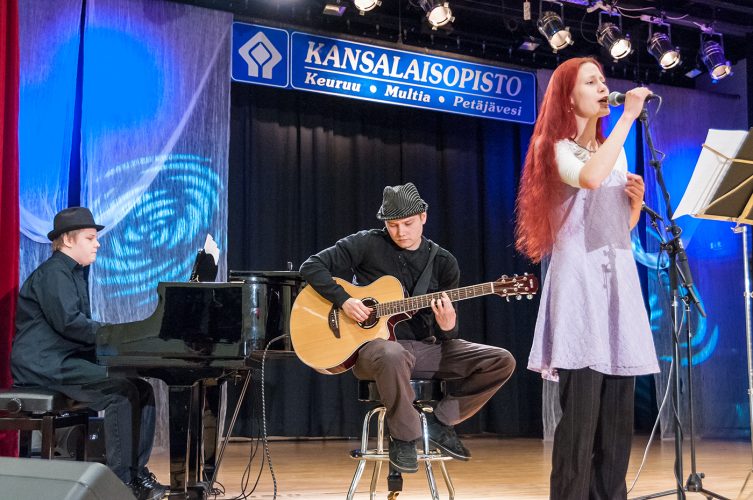 Pitkähiuksinen nainen laulaa mikrofoniin, taustalla kaksi mustahattuista miestä soittaa kitaraa ja pianoa.
