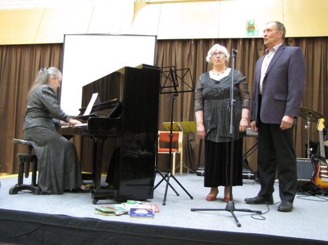 Mies ja nainen laulavat mikrofoniin, nainen säestää heitä pianolla.
